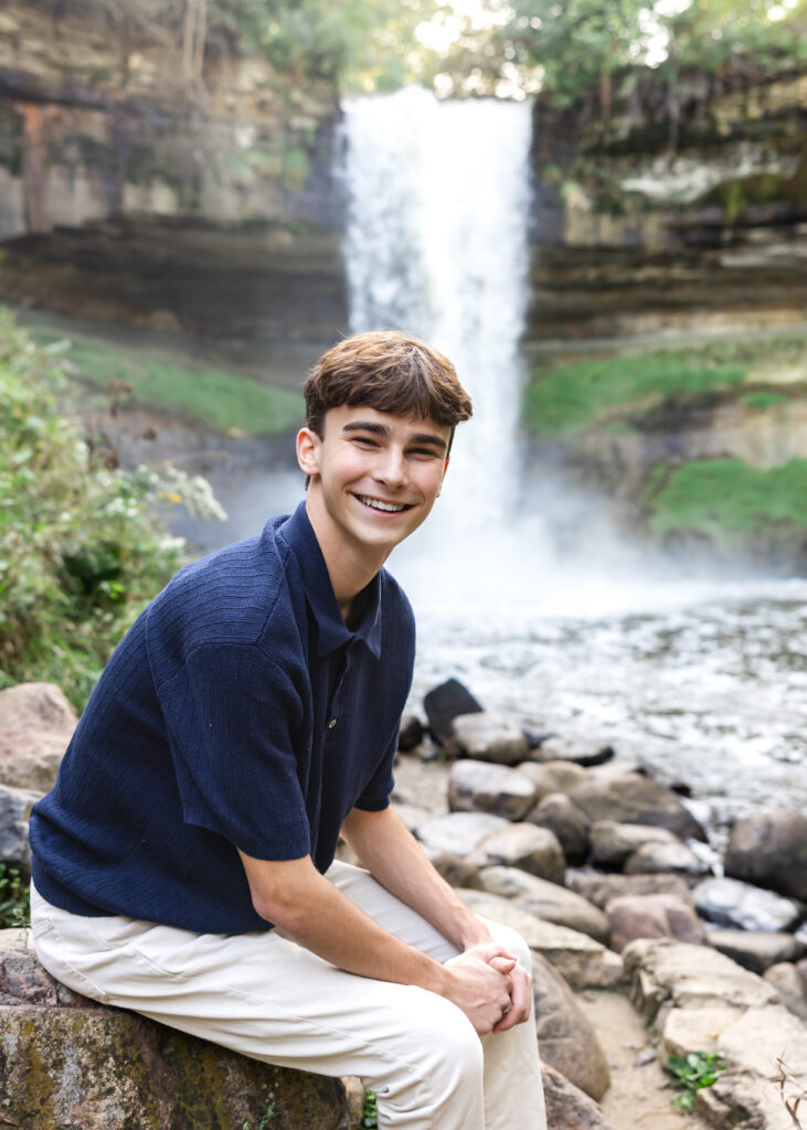 high school senior boy sits on a rock in front of minnehaha falls, minneapolis for his senior pictures session