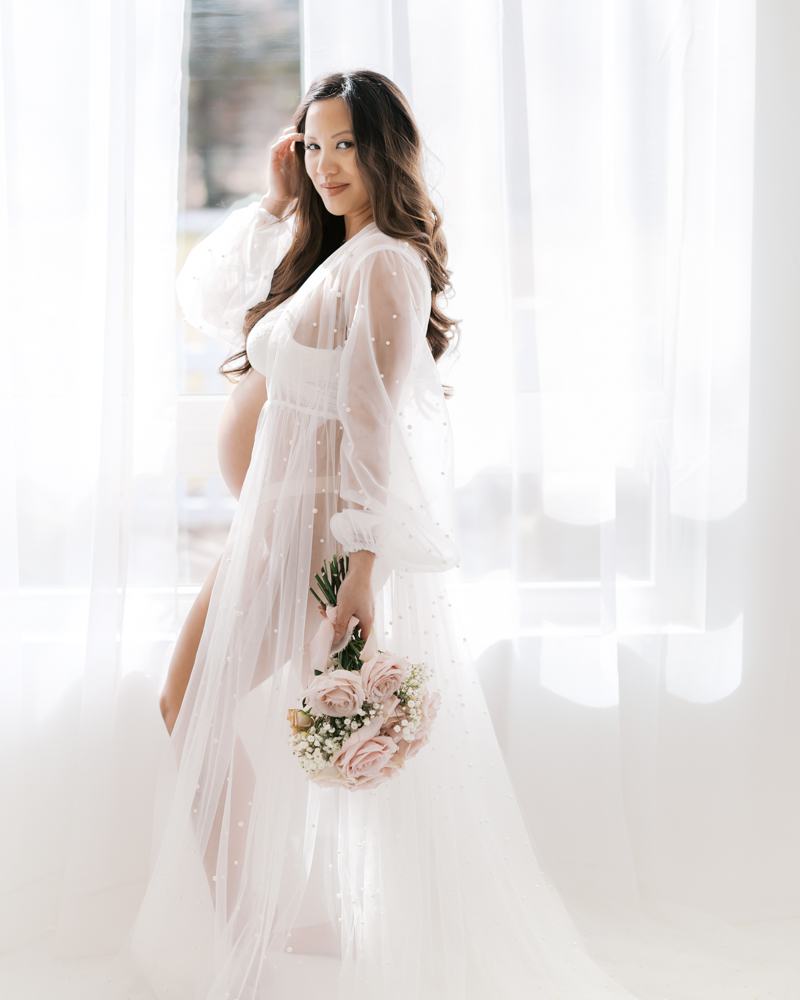 woman in long white maternity gown stands by a studio window and holds a bouquet of pink roses during her minneapolis maternity photoshoot