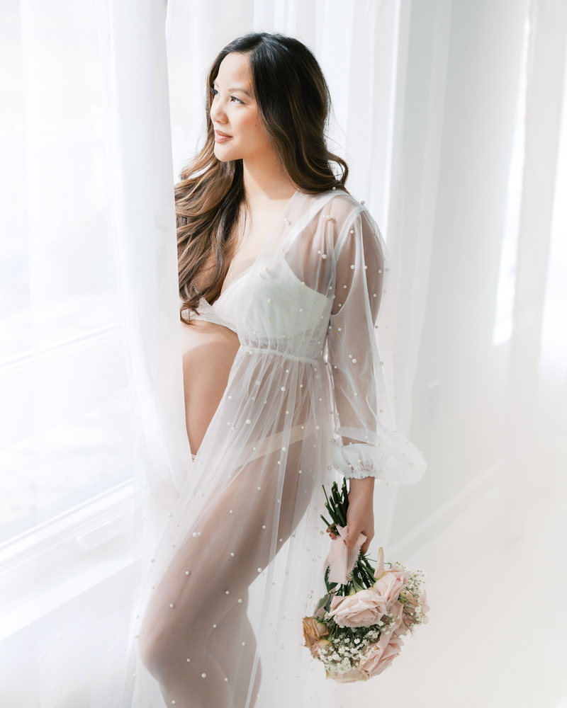 woman in sheer white maternity robe holds a bouquet of flowers and looks out a studio window during her minneapolis maternity photography session