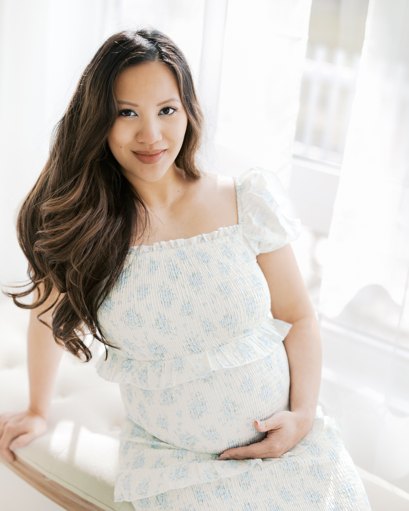 woman in blue and white floral dress holds her belly by a studio window during her minneapolis maternity photoshoot with angela watts