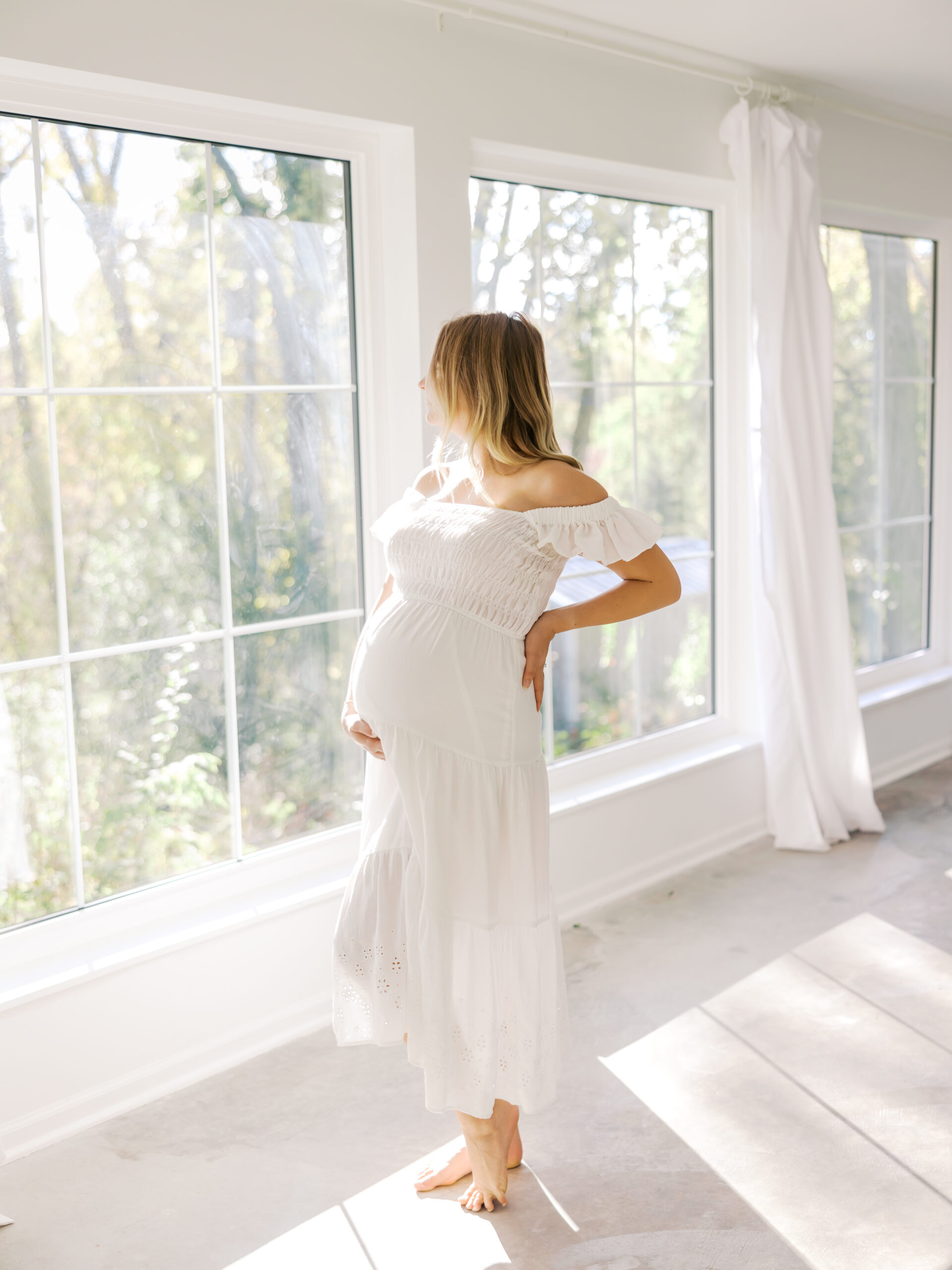 Woman in long white dress holds her pregnant belly during her in studio maternity photoshoot in minnetonka, mn with angela watts photography.