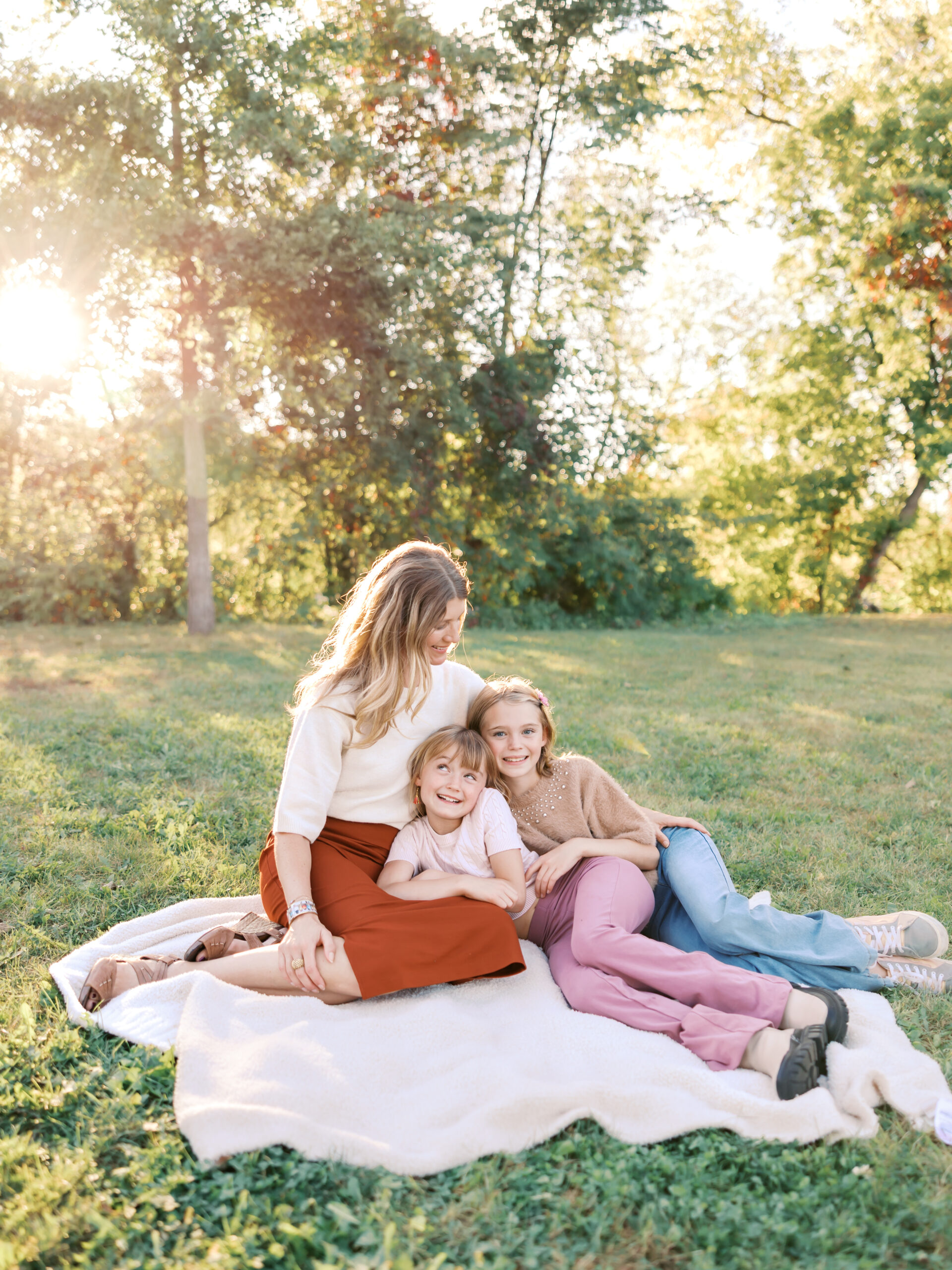 mom snuggles her two daughters on a blanket with sun shining behind at west medicine lake park in plymouth, mn for their minneapolis family photography session