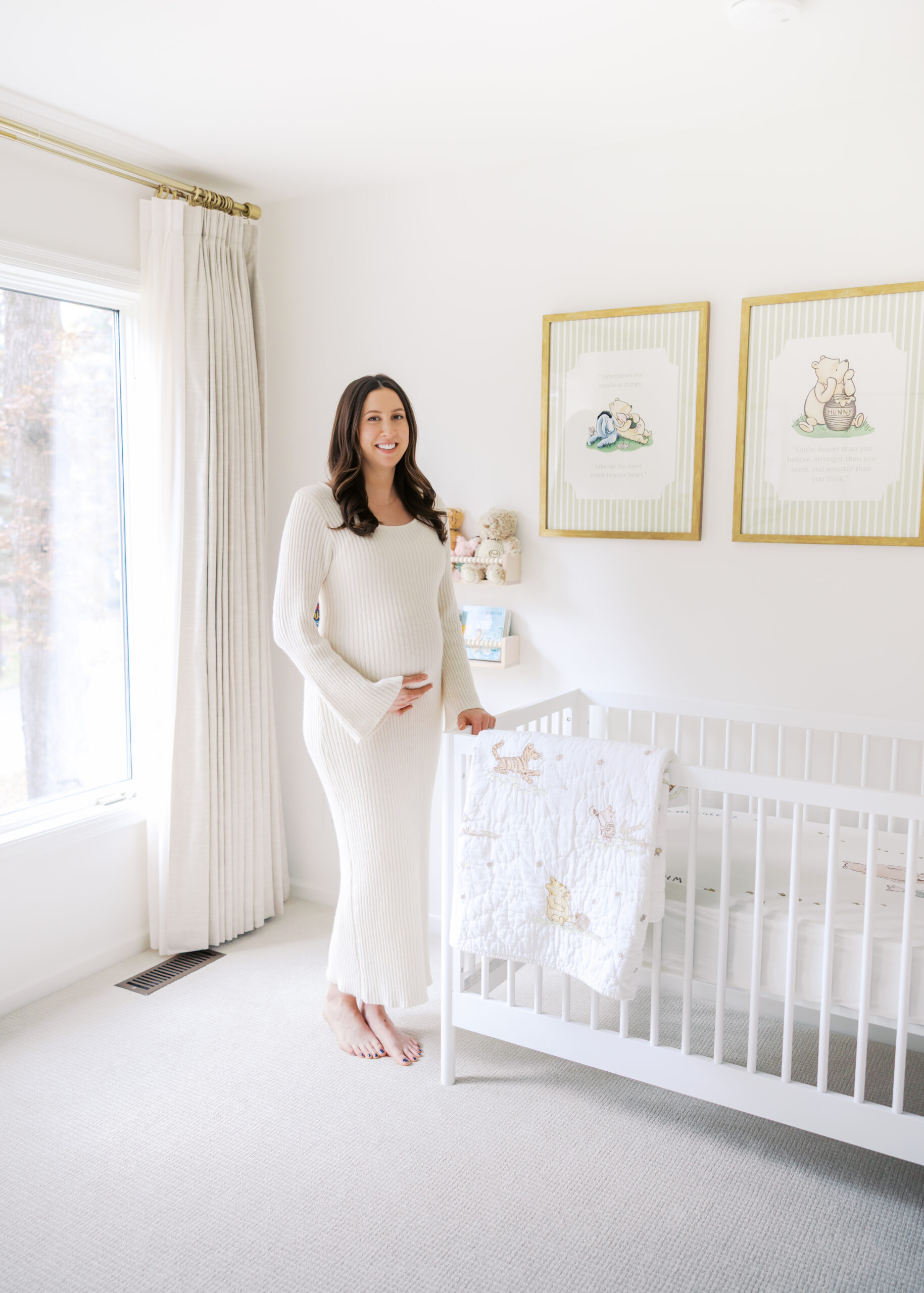Pregnant woman in white sweater dress holds belly while standing by crib in her in home minnetonka maternity photo session with angela watts photography