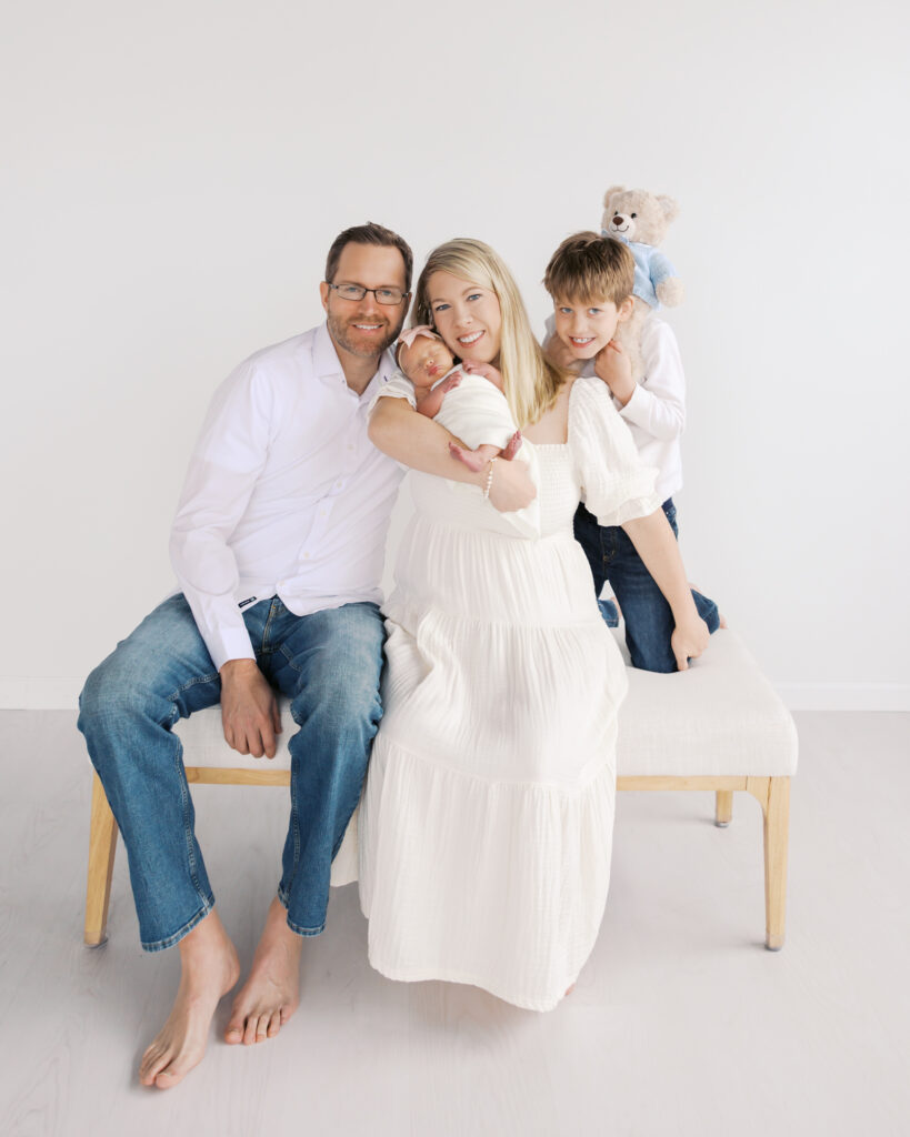 family of 4 sit on a bench in a studio setting during their minneapolis newborn photography session with angela watts