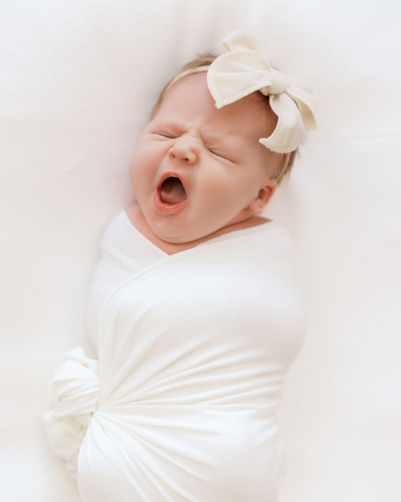 Newborn baby girl yawns while laying on white blanket during her newborn photography session in a minneapolis studio.