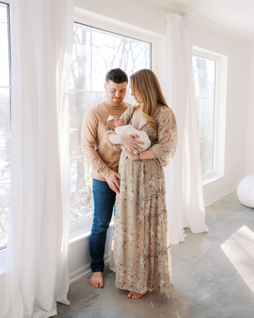 Mom dad and newborn baby girl stand in front of large windows in a light-filled minneapolis studio for their newborn photography session