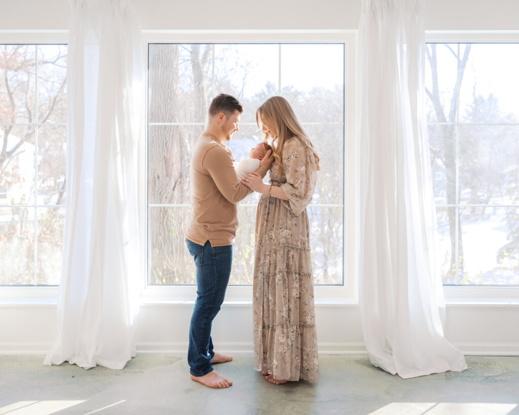 Mom and dad stand in front of large windows holding their newborn baby during a minneapolis studio newborn session.