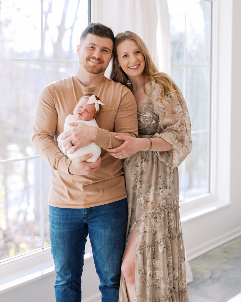 Mom dad and newborn baby girl stand in front of large windows in a light-filled minneapolis studio for their newborn photography session