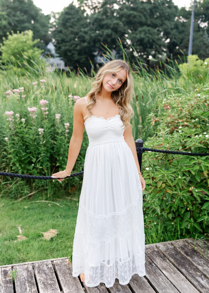 Girl in long white dress poses at wayzata beach for her wayzata senior pictures with angela watts photography