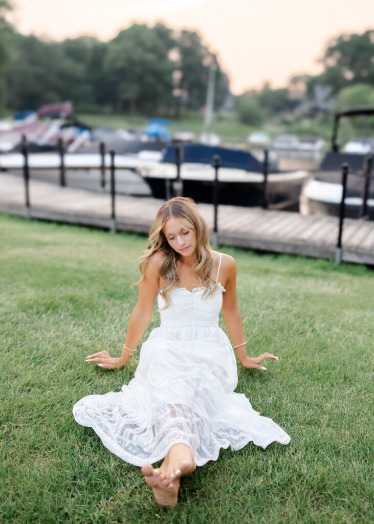 Girl in long white dress poses at wayzata beach for her wayzata senior pictures with angela watts photography