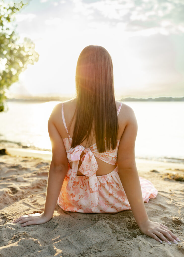 Girl sits at golden hour at Excelsior beach in white and pink flowy dress for her eden prairie senior pictures with senior photographer angela watts photography.