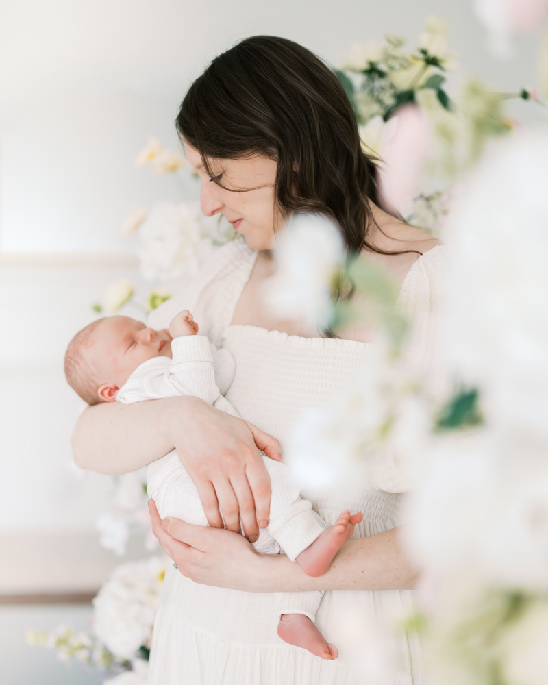 mom holds newborn baby boy surrounded by flowers in a studio setting for her minneapolis newborn photography session with angela watts.