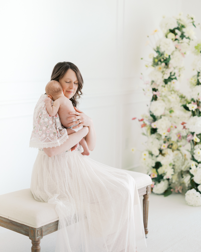 mom in white lacy dress holds newborn baby boy in her arms in a studio setting during their minneapolis newborn photography session