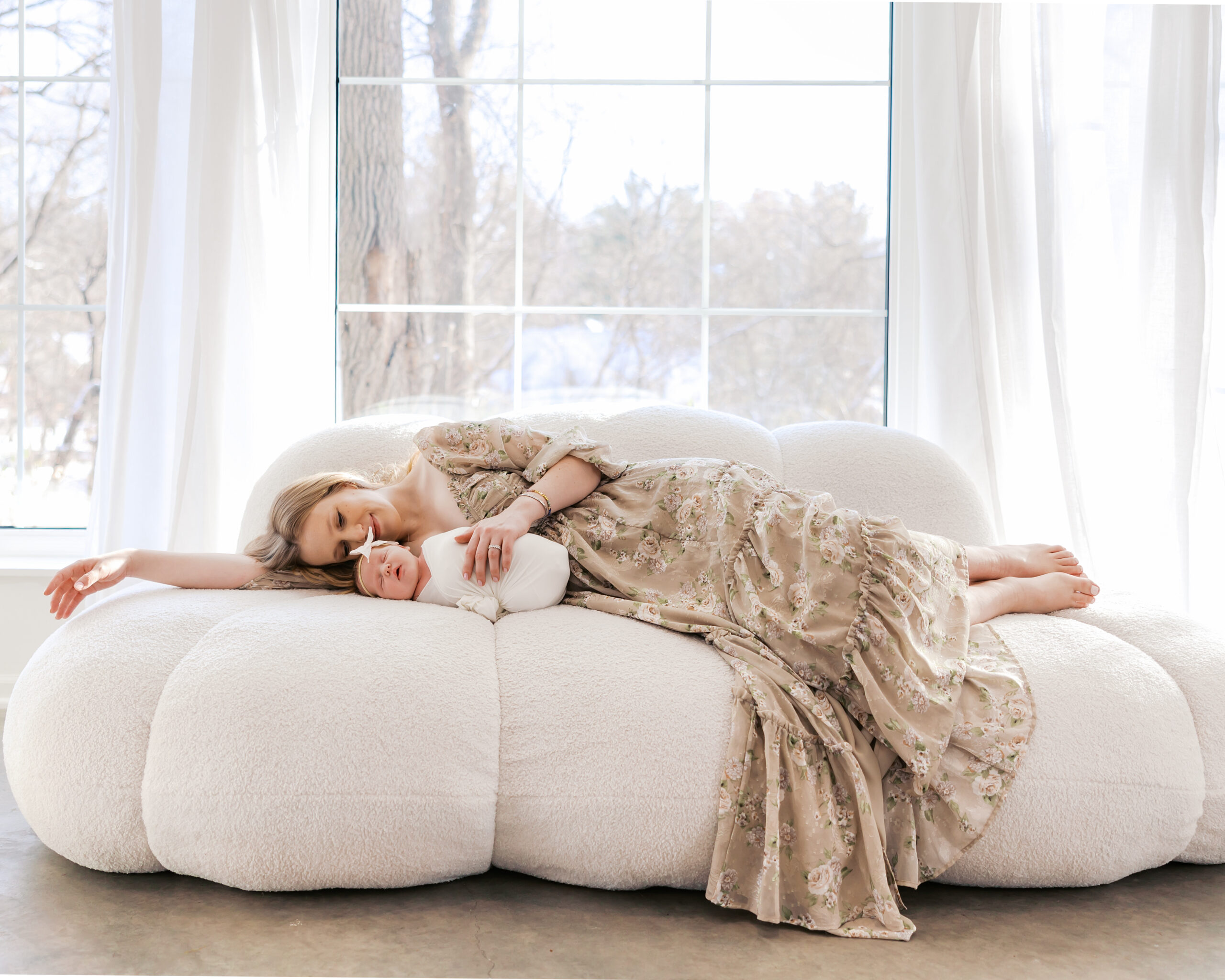 Mom and baby lie on a sofa in front of huge bright windows in a light-filled minneapolis studio during their newborn photography session with angela watts photography