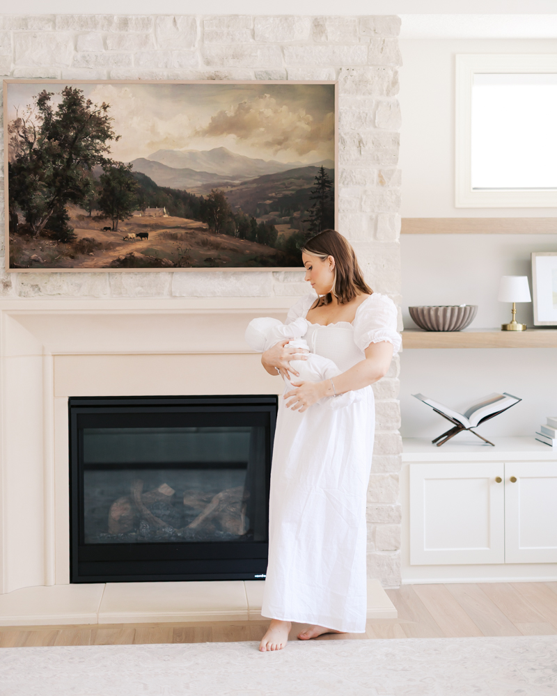 Woman in white dress holds newborn baby in their edina living room during their newborn photos with edina newborn photographer angela watts photography.