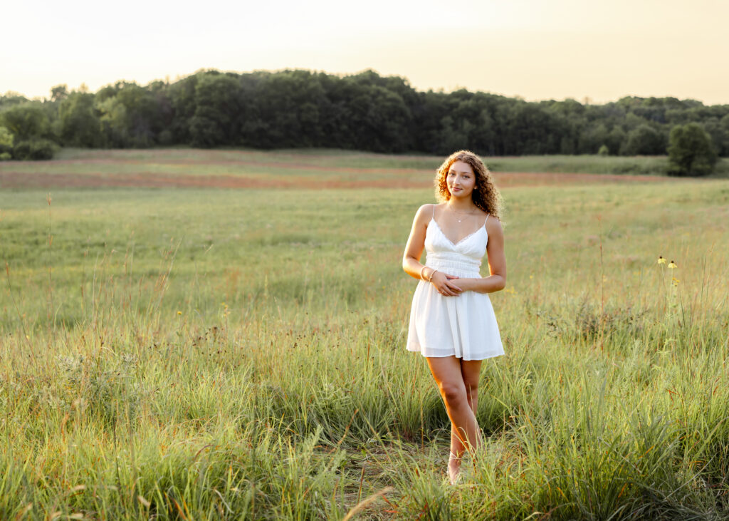 girl wearing white dress stands in a dreamy field at richardson nature center during her bloomington senior photos with angela watts photography
