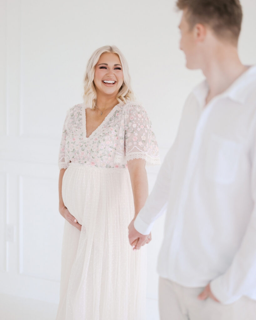 husband holds pregnant wife's hand in a light-filled studio for their minneapolis maternity photo session with angela watts photography