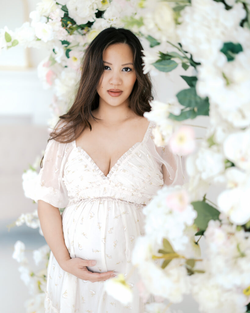 woman in pale cream dress holds her belly surrounded by flowers in a studio setting for her minneapolis maternity photoshoot