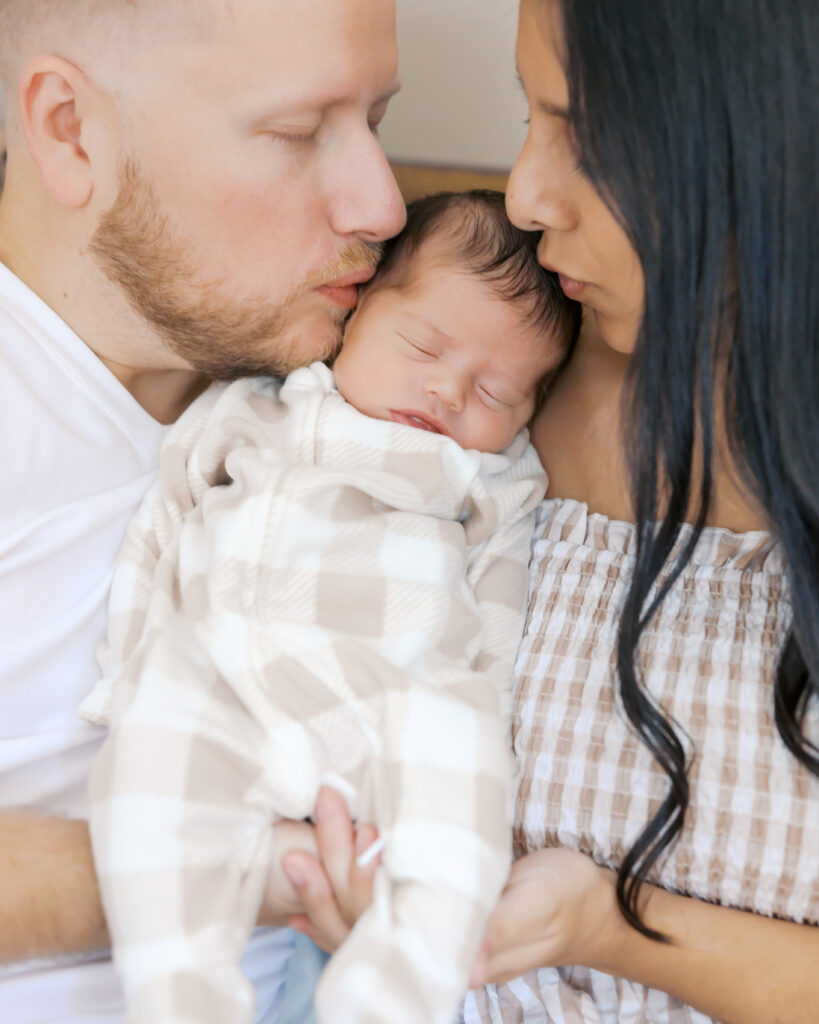 2 year old girl with pigtails sits on her parents bed while parents snuggle their newborn in the background during their edina, minnesota newborn photography session