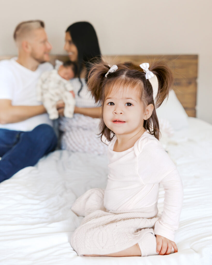 2 year old girl with pigtails sits on her parents bed while parents snuggle their newborn in the background during their edina, minnesota newborn photography session