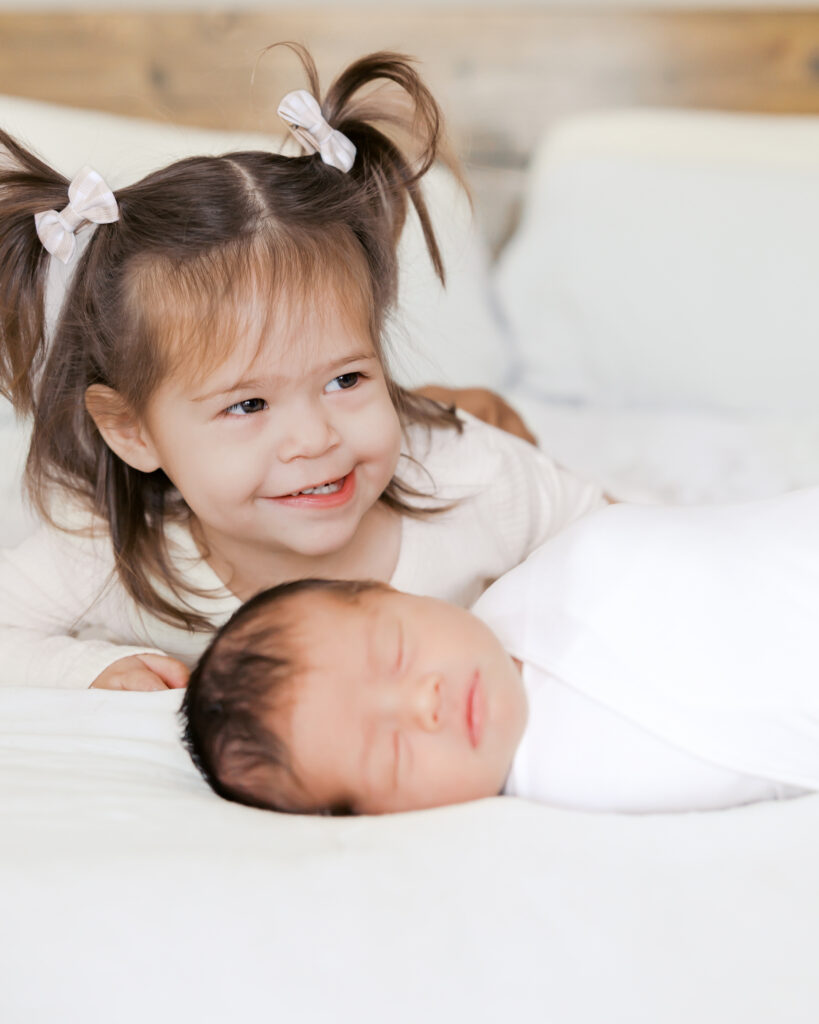 2 year old girl with pigtails sits on her parents bed while parents snuggle their newborn in the background during their edina, minnesota newborn photography session