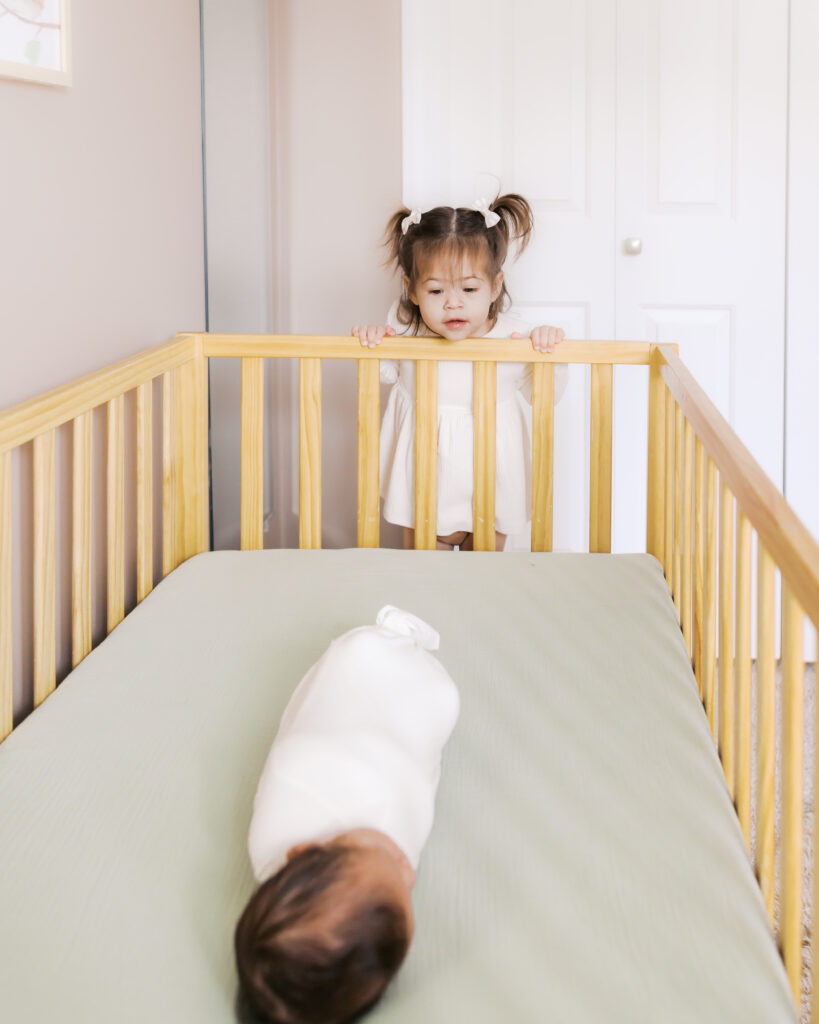 2 year old sister peeks over crib with newborn brother sleeping inside in their edina newborn photography session