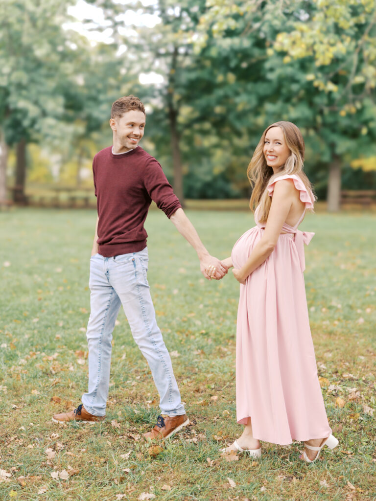 pregnant woman wearing pink ruffled dress holds husband's hand in a minneapolis park for their minneapolis maternity photos
