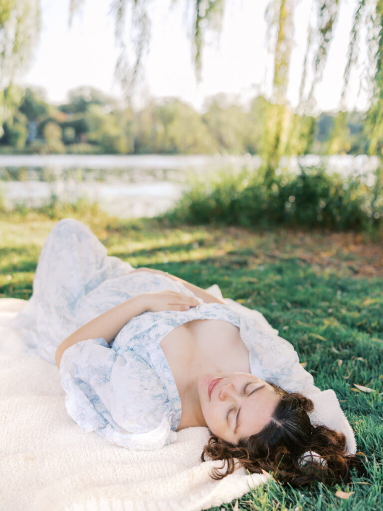 woman in light blue and white dress lies in the grass at lake of the isles in minneapolis for her maternity photoshoot