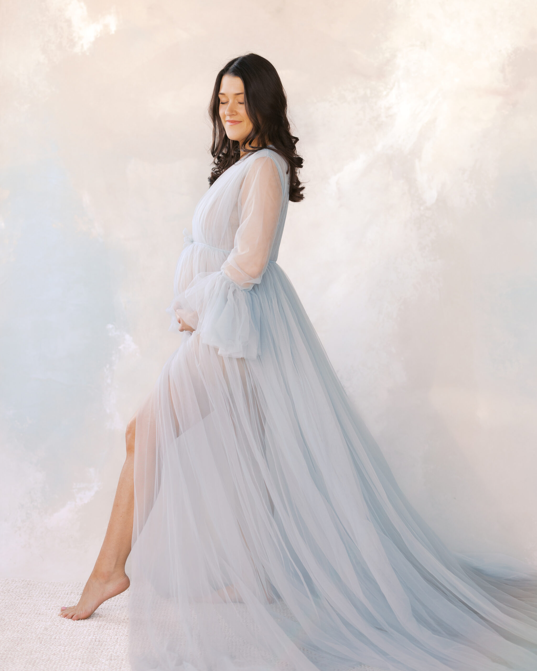 woman in long elegant, blue maternity gown poses in front of a serene studio backdrop for her minneapolis maternity photos.