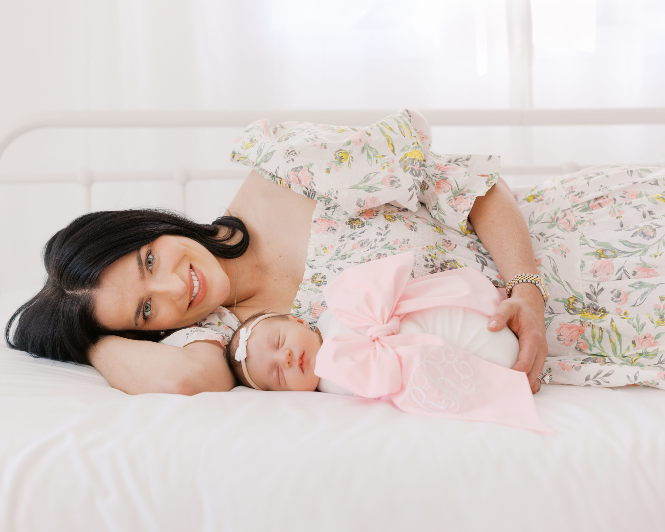 elegant woman in pink and white dress lies on a bed with her newborn baby girl