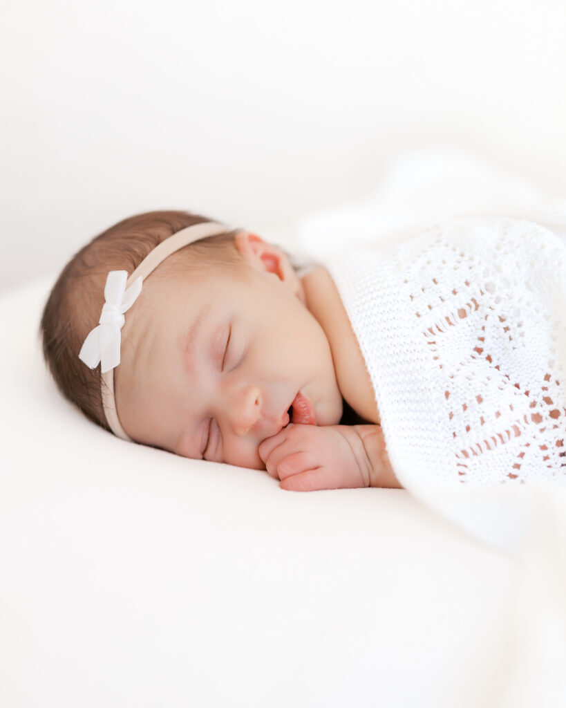 Newborn baby girl with white bow sleeps on a white bean bag covered in a detailed white blanket during her newborn photography session.
