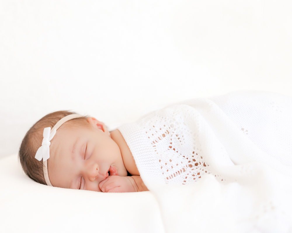 Newborn baby girl lying on white bean bag covered in white blanket sleeps during her minneapolis newborn photography session in a light-filled studio setting.