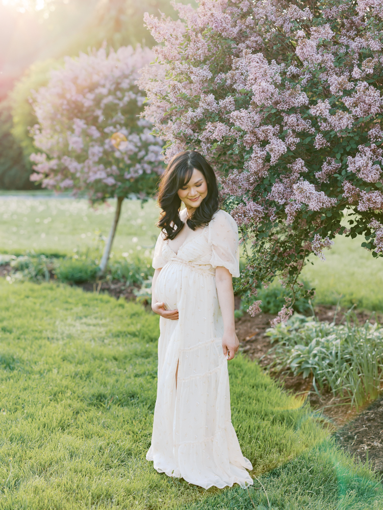 woman wearing long cream colored dress stands in front of lilac trees at lyndale park rose garden for her minneapolis maternity photography session