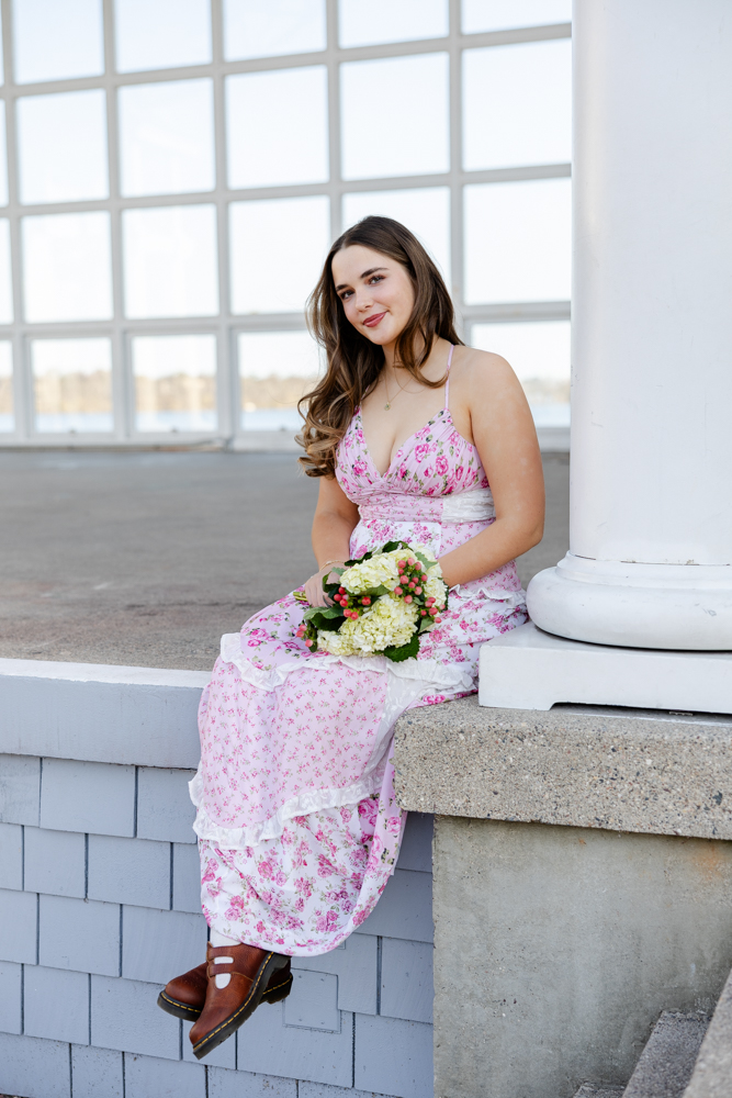 girl in long pink dress sits at lake harriet bandshell holding a bouquet for her minneapolis senior photography session.