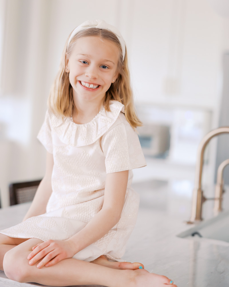 girl in white ruffly dress and white headband sits on kitchen counter of her edina, minnesota home for her edina family photography session
