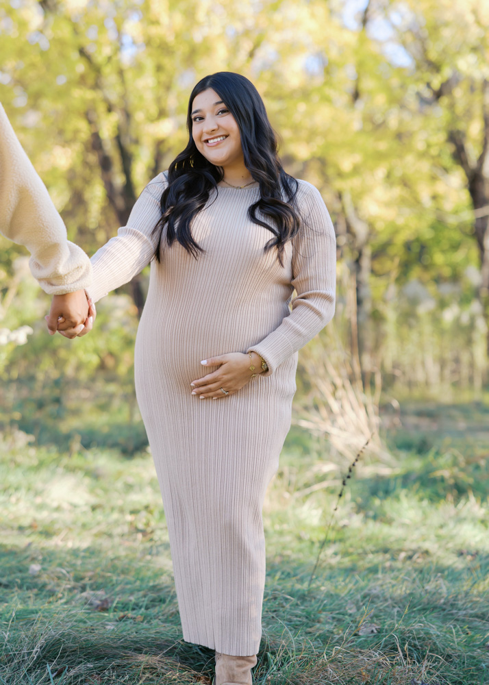woman in beige sweater dress holds her pregnant belly at longfellow gardens for her minneapolis maternity photos with angela watts