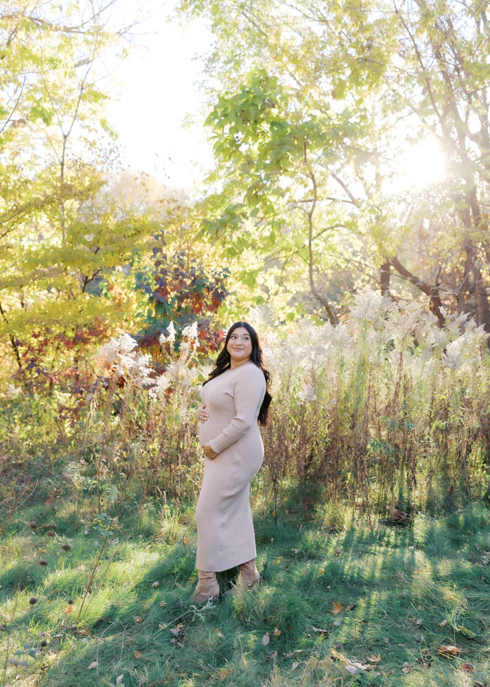 woman in beige sweater dress holds her pregnant belly at longfellow gardens for her minneapolis maternity photos with angela watts