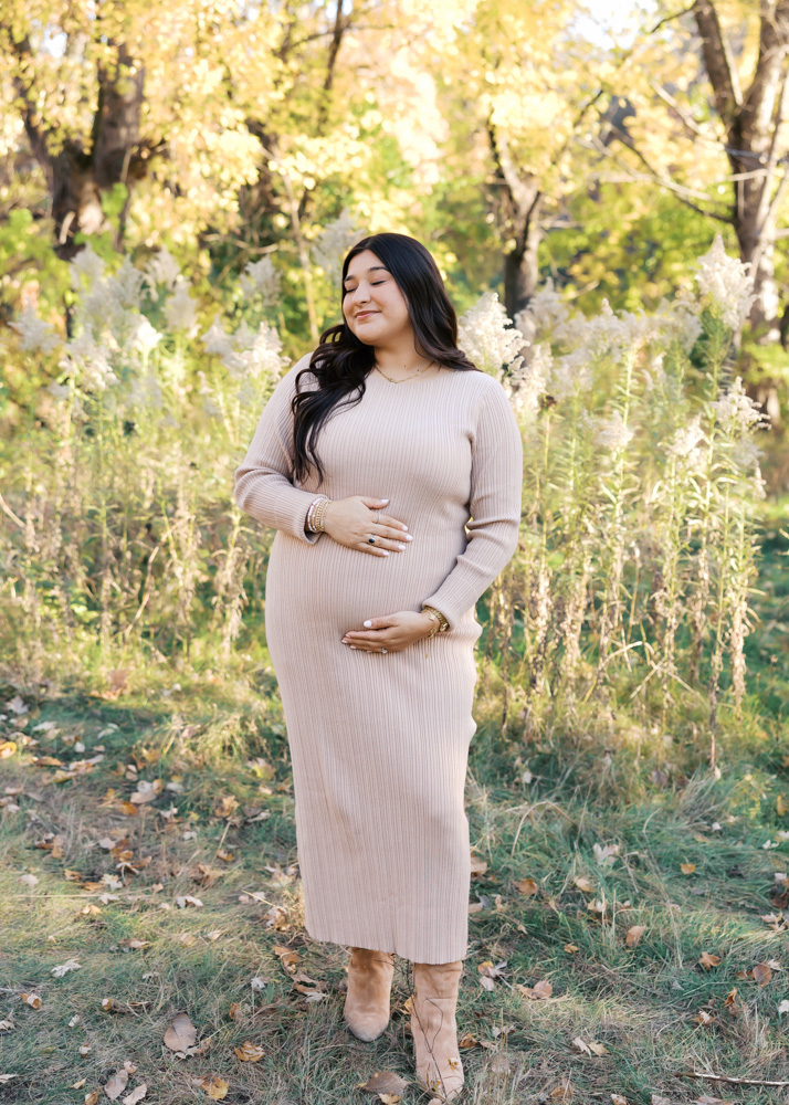 woman in beige sweater dress holds her pregnant belly at longfellow gardens for her minneapolis maternity photos with angela watts