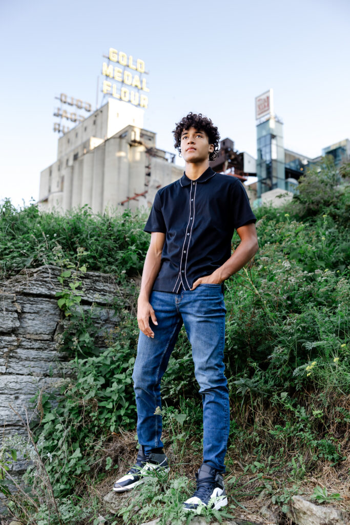 boy in black shirt and jeans poses at mill ruins park for his minneapolis senior pictures