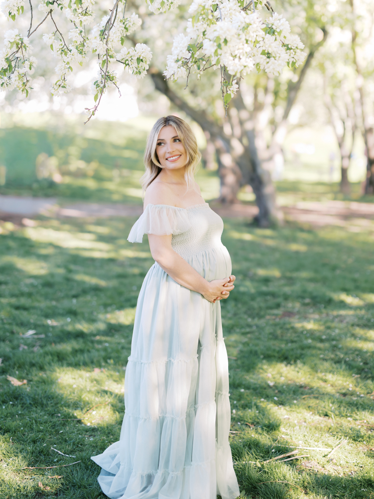 pregnant woman in mint green tulle gown stands under the white apple blossoms during her minneapolis maternity photoshoot with angela watts