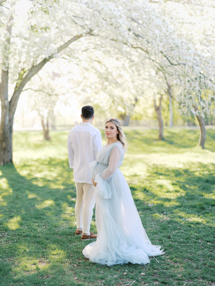 Husband and wife hold hands under the big white apple blossoms at lyndale park rose garden during their minneapolis maternity photography session