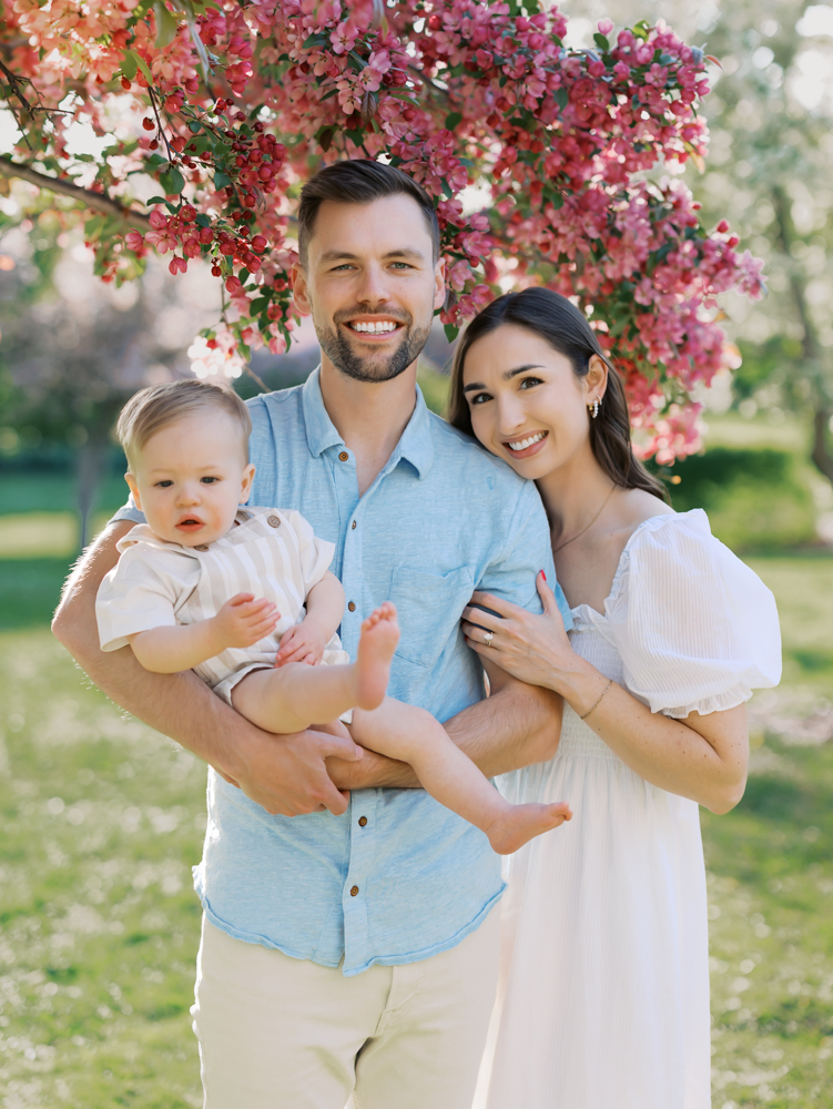 family of 3 wearing blue and white stands in front of beautiful pink apple blossoms at lyndale park gardens for their minneapolis family photos