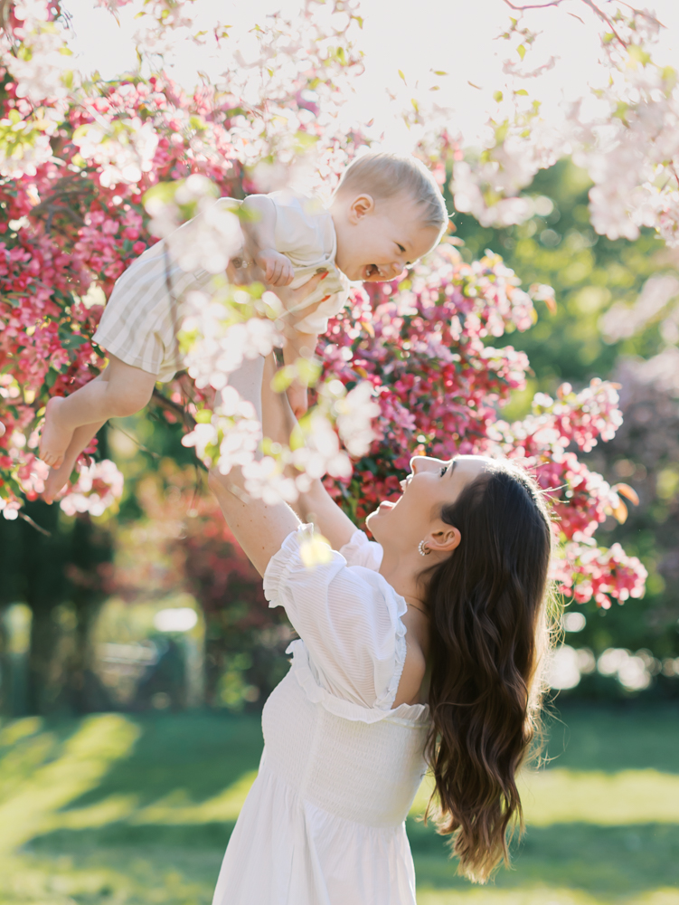 mom holds up 14 month old baby boy in the middle of white and pink apple blossoms at lyndale park gardens for their minneapolis family photography session