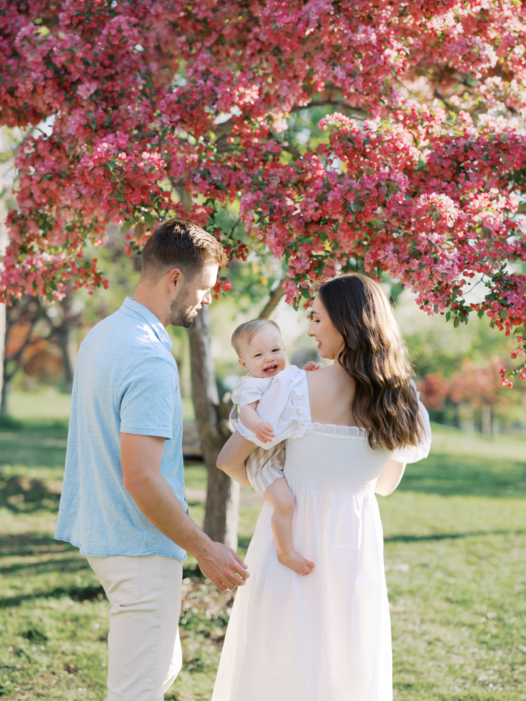 family of three wearing blue and white with one year old boy standing under pink crabapple blossoms during their minneapolis family photography session
