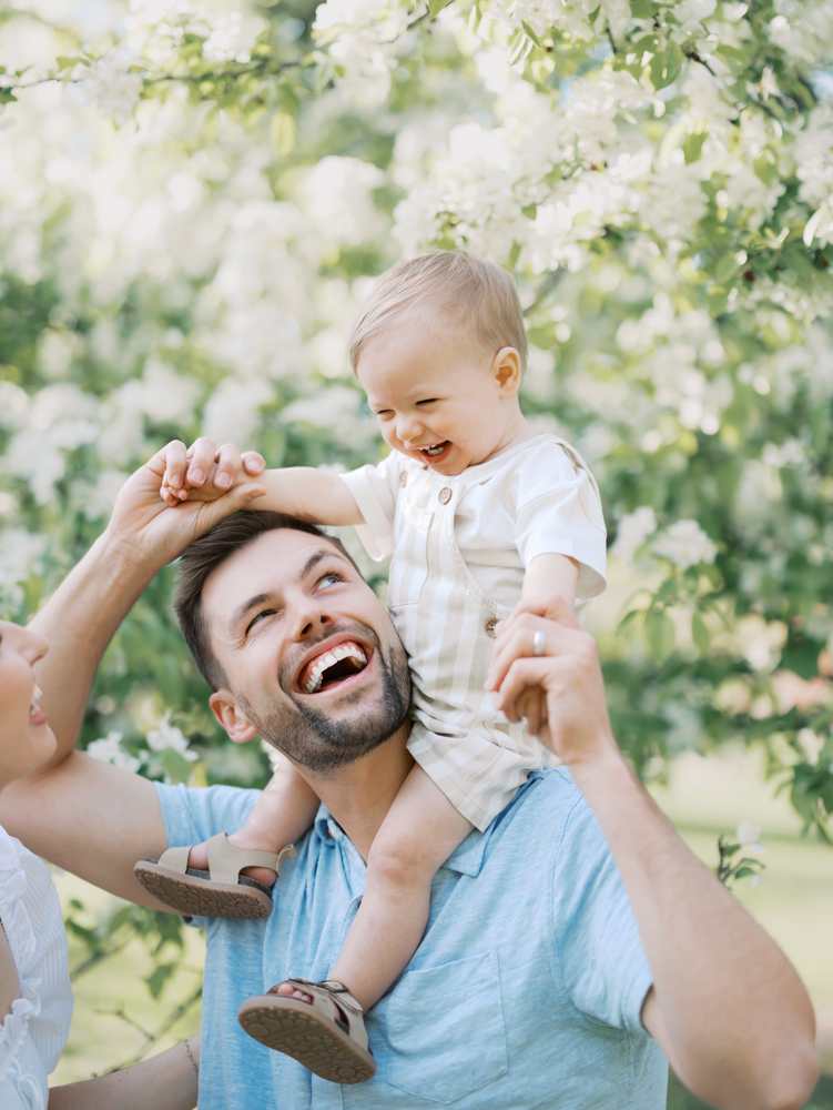 dad holds baby boy on his shoulders in front of white apple blossoms at lyndale park gardens for their minneapolis family photos