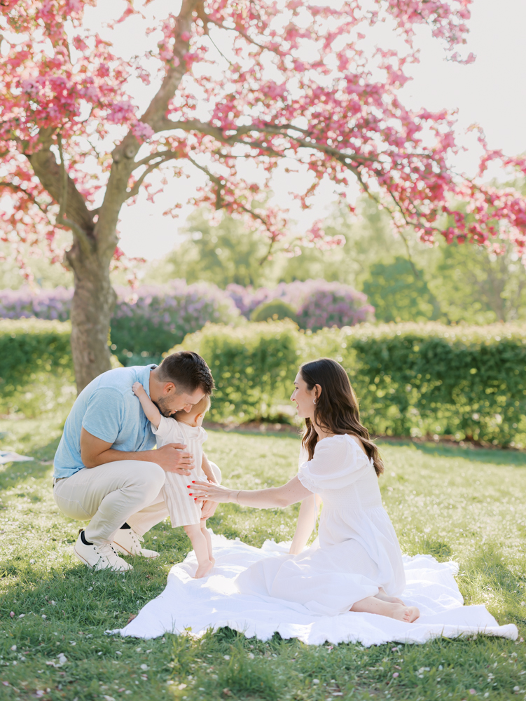 mom, dad and baby sit on a blanket in front of a pretty pink tree at lyndale park gardens for their minneapolis family photography session