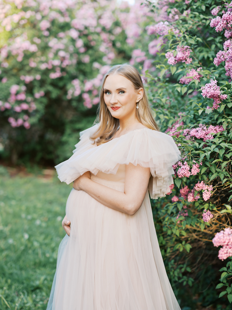 woman in peach tulle maternity gown stands in front of large lilac bushes at mn landscape arboretum for her minneapolis maternity photoshoot