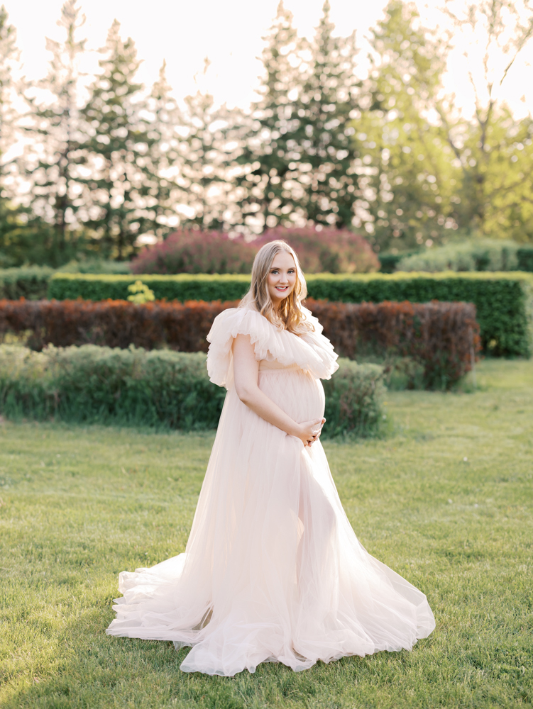 woman in blush colored maternity gown stands near hedges at mn landscape arboretum for her minneapolis maternity photography session